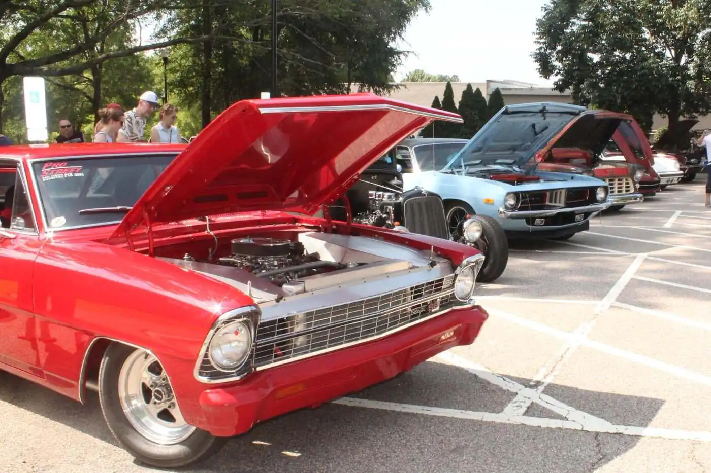 A row of show cars with their hoods raised, led by a red Chevrolet Nova pro-street build, as spectators walk the field