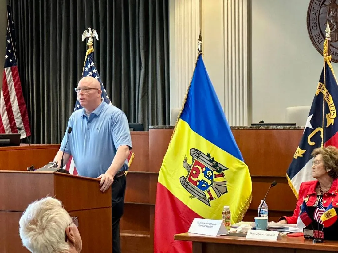 Speaker at a podium beside the Moldovan and North Carolina flags, with Secretary of State Elaine F. Marshall seated at right