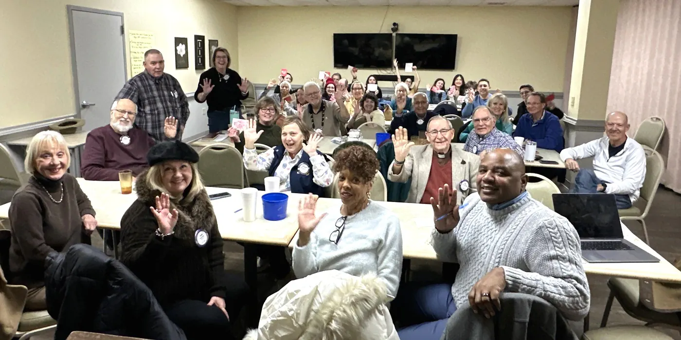 Members of the Rotary Club of Wake Forest seated at dinner tables, waving at the camera during a Monday evening meeting