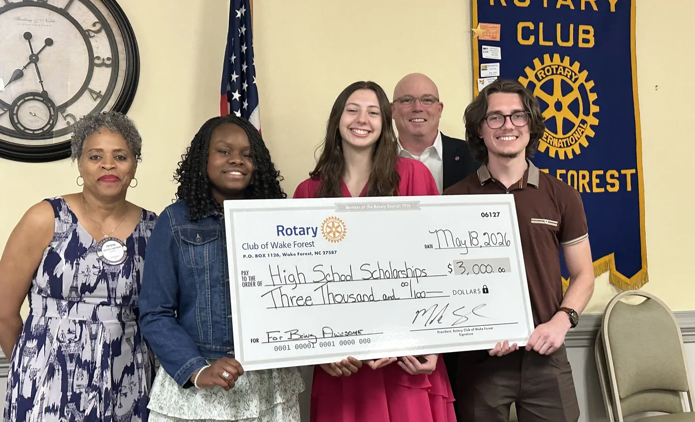 Three graduating seniors hold an oversized $3,000 High School Scholarships check with two Rotary Club of Wake Forest members at a club meeting
